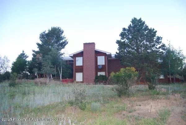 Two-story brick house with a central chimney, surrounded by pine trees and shrubs.