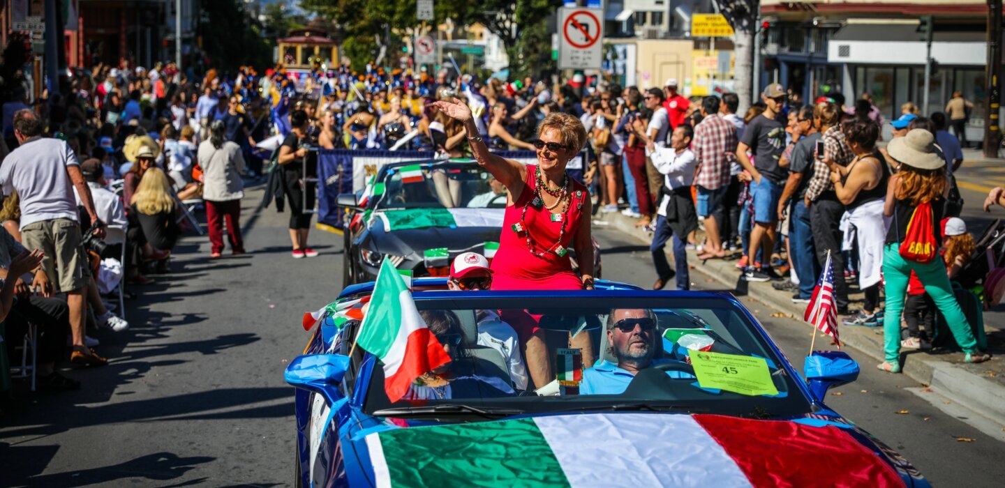 A lively parade scene in North Beach, San Francisco, featuring a woman in a red dress waving from a blue convertible decorated with the Italian flag. The street is packed with spectators, many holding flags and cheering, while a marching band and cable car can be seen in the background. The celebration appears to be an Italian heritage event, with vibrant colors and festive energy filling the scene.