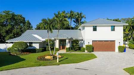 White two-story home with a gray metal roof, brown garage door, and palm trees along the lawn.