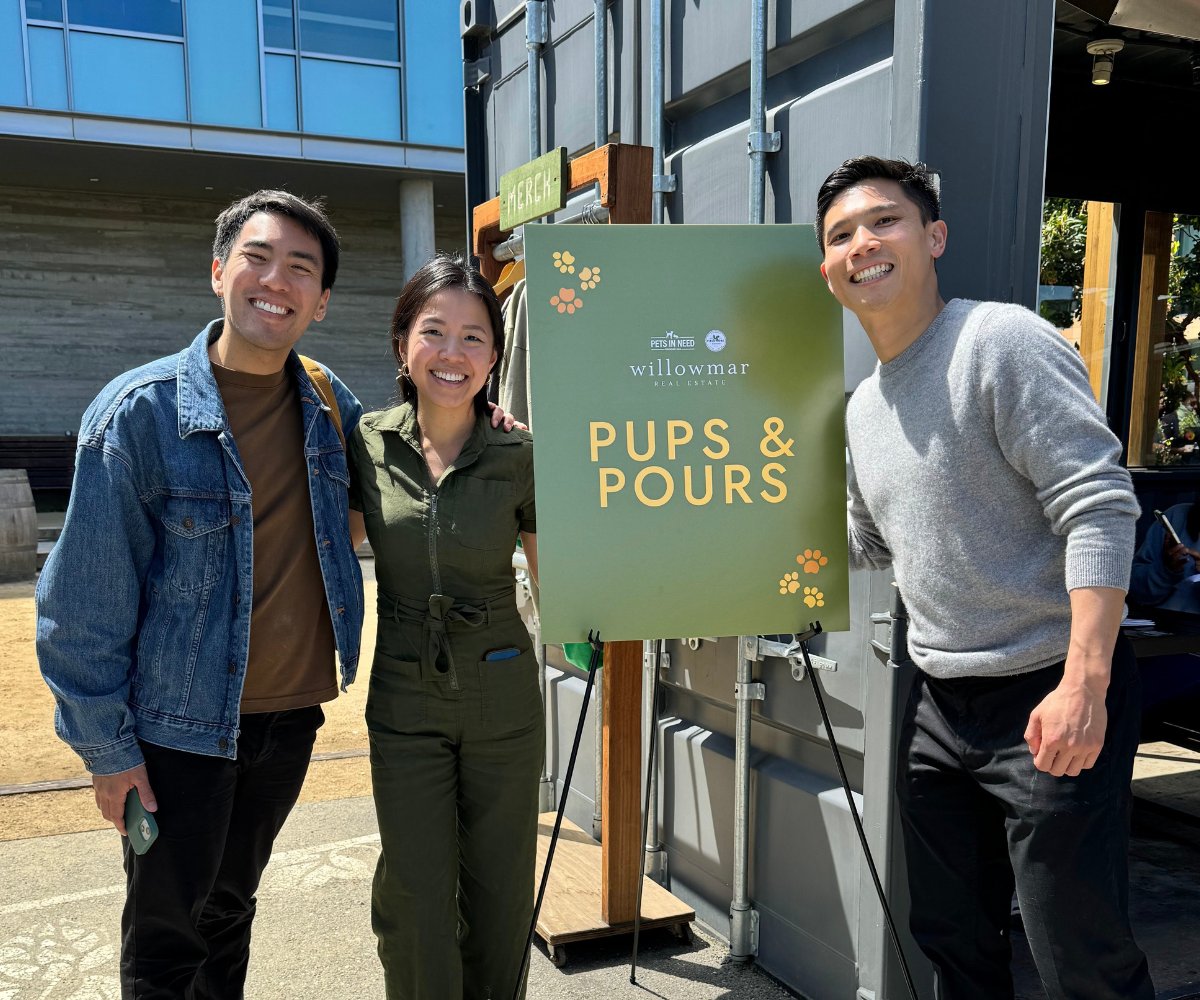 Three Willowmar Real Estate colleagues pose beside a green “Pups & Pours” event sign.