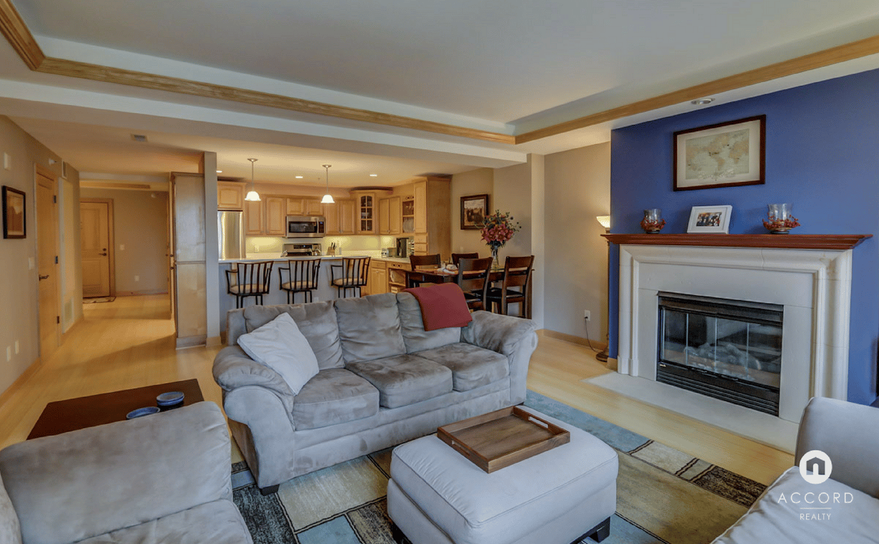 Condo living room with gray sectional, white fireplace, and open kitchen.