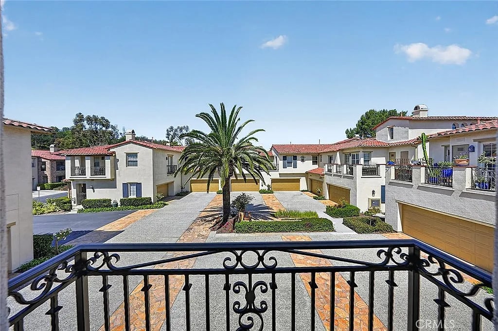 Townhome complex with red tile roofs and a central palm, seen over an ornate balcony railing.