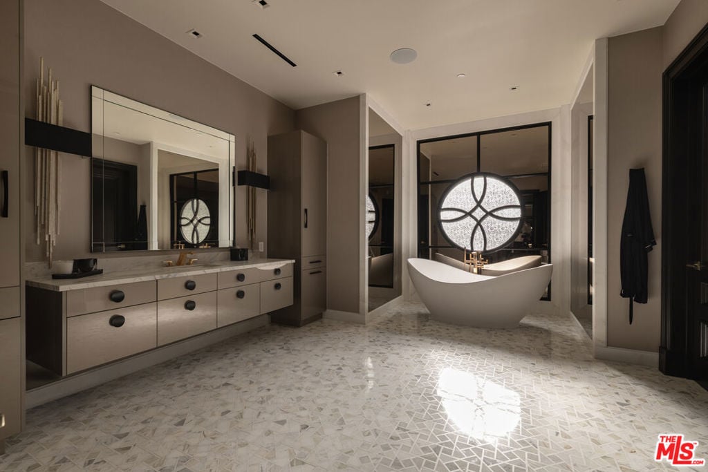 Luxury bathroom with a freestanding tub, circular window, double vanity, and geometric stone tile floor.