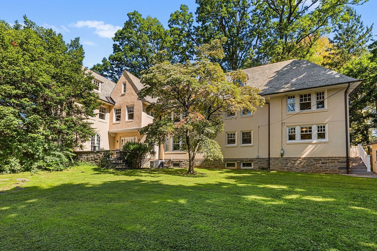 Large beige house with a stone foundation, gray shingle roof, and broad lawn shaded by mature trees.