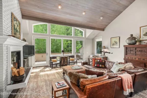 Living room with a vaulted wood ceiling, white brick fireplace, and large windows overlooking greenery.
