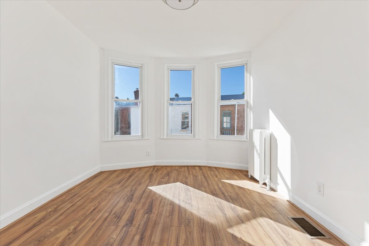 Vacant room with three tall windows, hardwood flooring, and a radiator.
