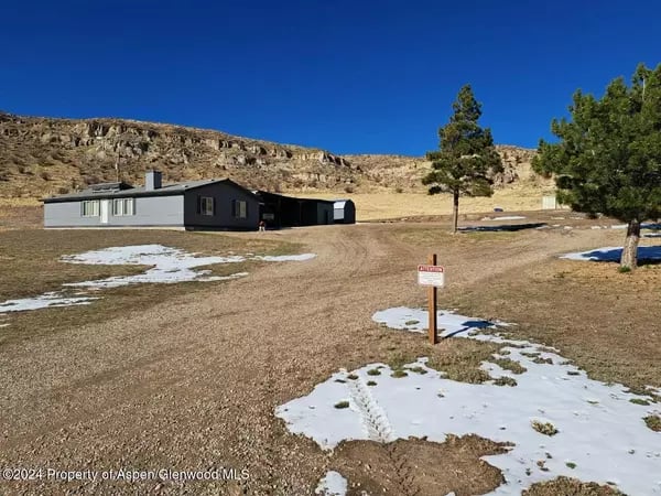 Single-story home on a broad dirt lot with patchy snow, backed by a rocky ridge.