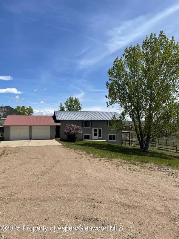 Gray two-story home with an attached garage, gravel driveway, and large leafy tree.