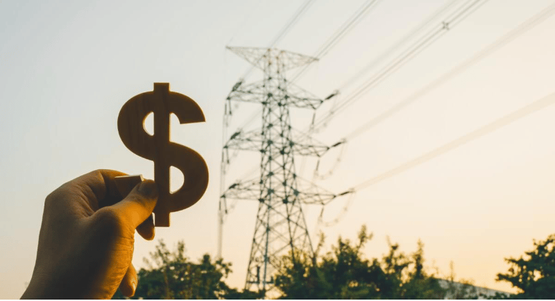 A hand holds a dollar sign in front of an electricity pylon at sunset.
