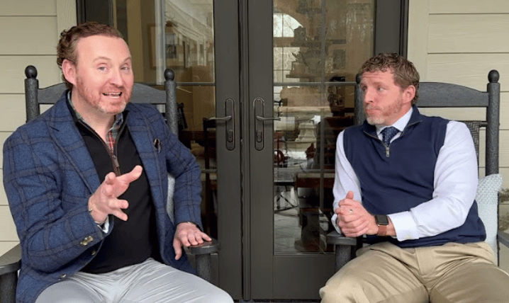 Two men seated outside glass doors, one gesturing as the other listens.