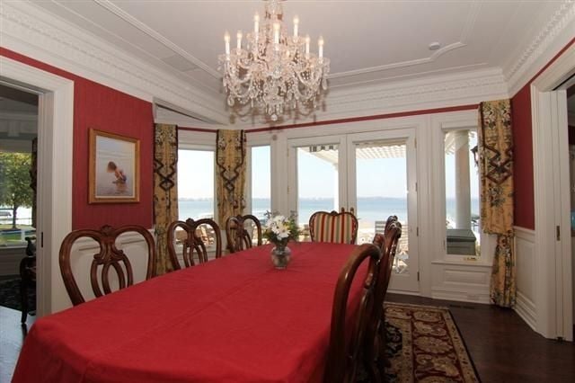 Formal dining room with a red-covered table, crystal chandelier, and lake views through tall windows.