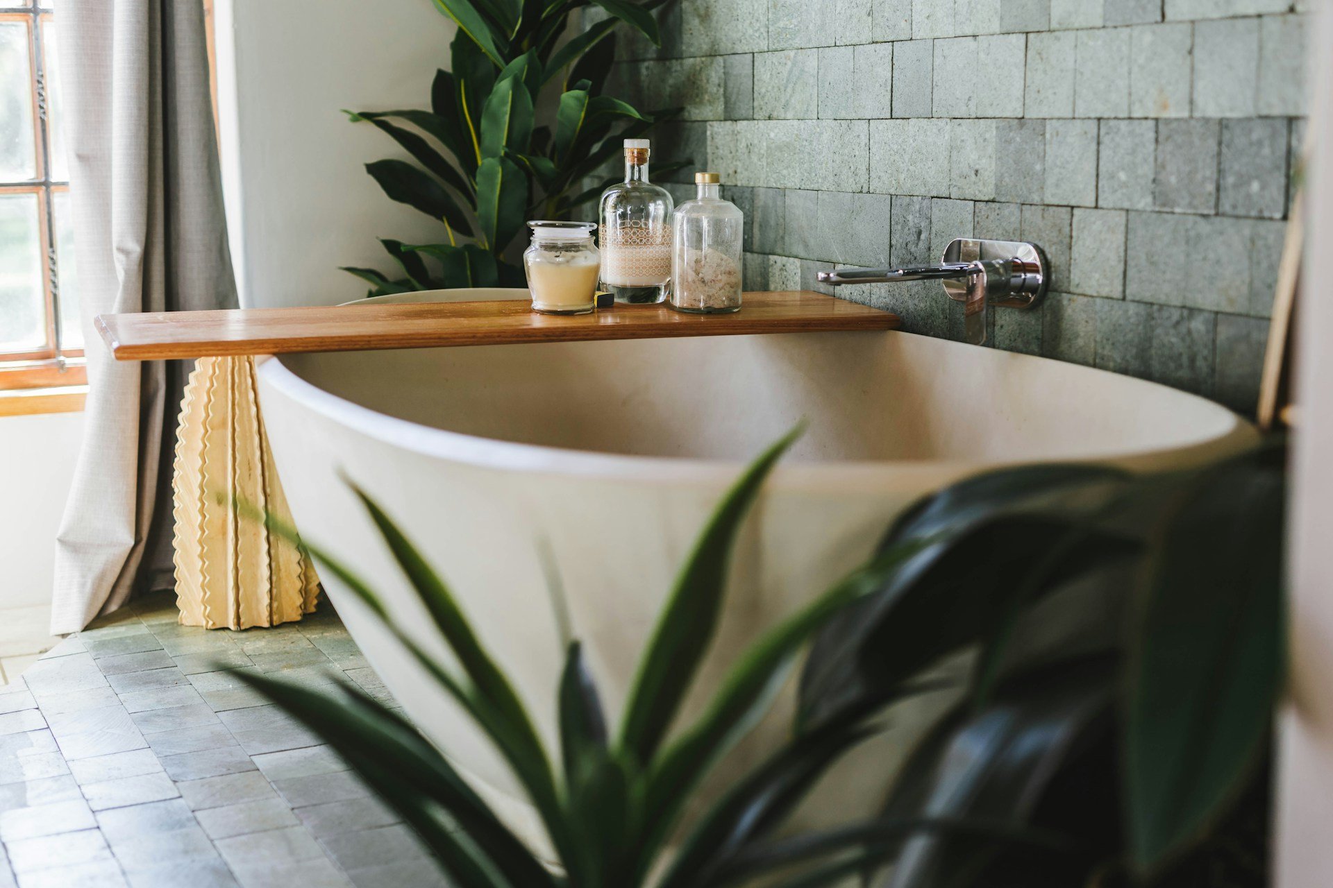 Freestanding bathtub with a wooden shelf, stone-tile wall, and leafy plants.