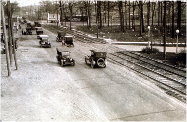 Early automobiles travel beside streetcar tracks on a tree-lined city street.