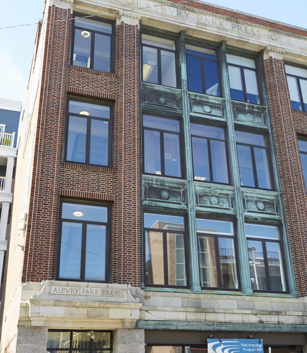 Brick building facade with large windows, ornate green metalwork, and Asbury Park Press stone signage.