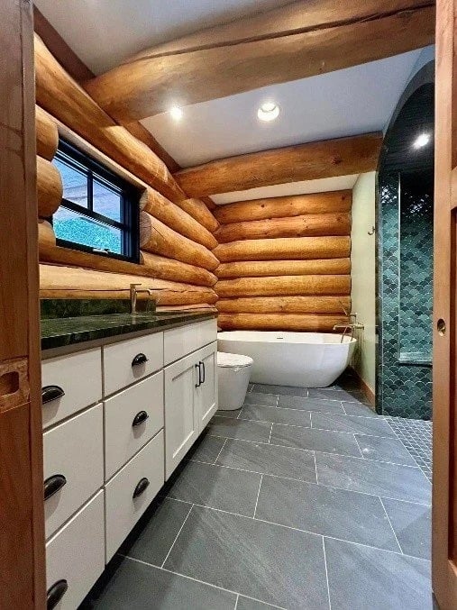 Log home bathroom with exposed timber walls, a freestanding tub, white vanity, and blue-green tiled shower.