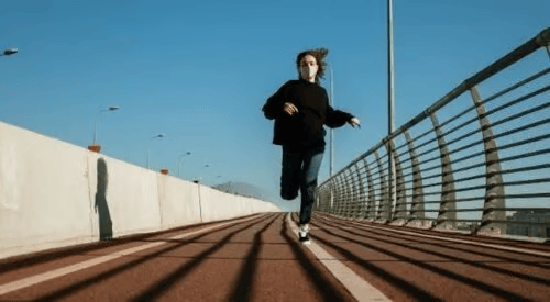 Masked runner on a pedestrian bridge with long shadows across the walkway.