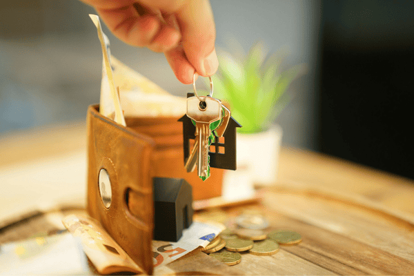 A hand holds house keys above a wallet, cash, and coins.