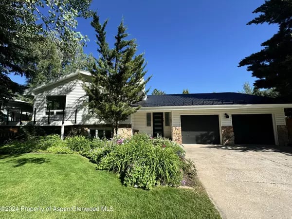 Front of a two-story home with an attached garage, lawn, shrubs, and mature evergreen trees.