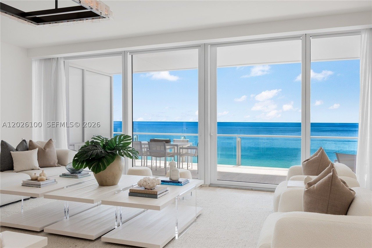 Staged oceanfront living room with white sofas and sliding doors opening to a balcony overlooking the sea.