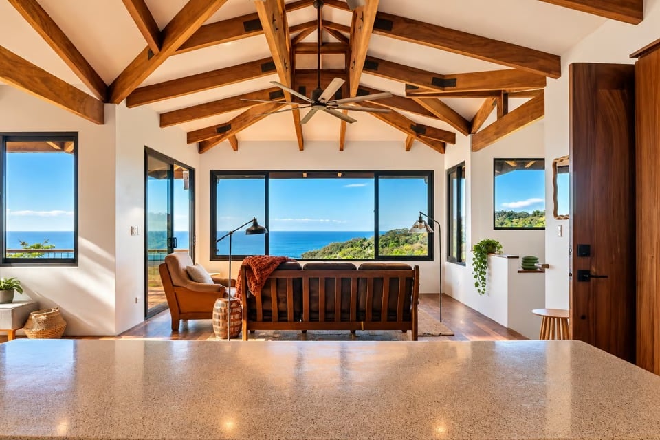 Furnished living room with exposed timber beams and broad windows overlooking the ocean and green hills.