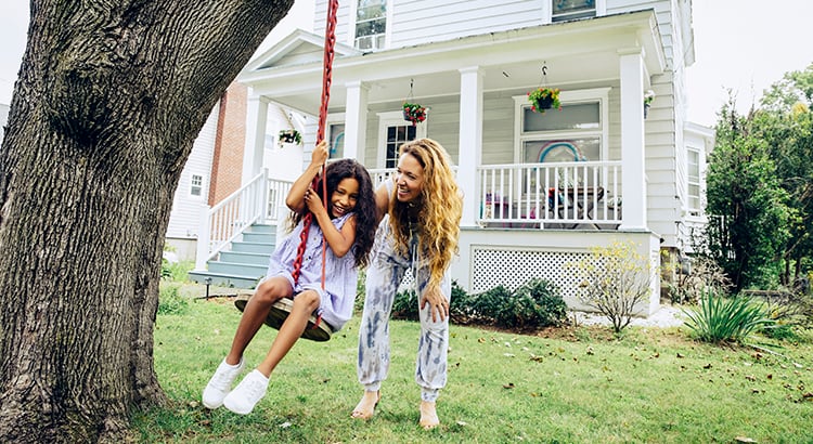 White two-story house with a covered porch behind a woman and girl on a tree swing.
