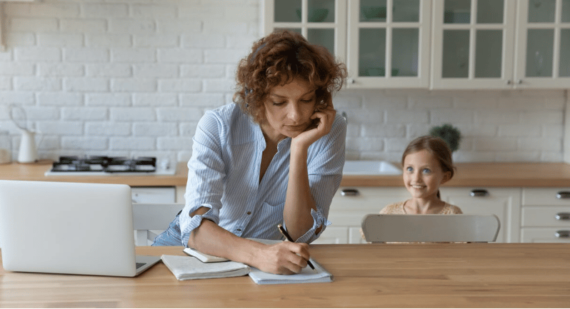 A mother writes at a kitchen table while a young girl sits behind a laptop.