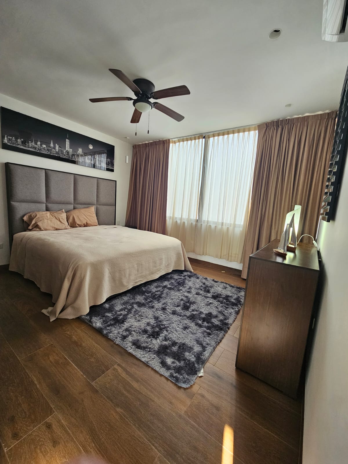 Bedroom with a large bed, upholstered headboard, patterned rug, and tall curtains over a bright window.