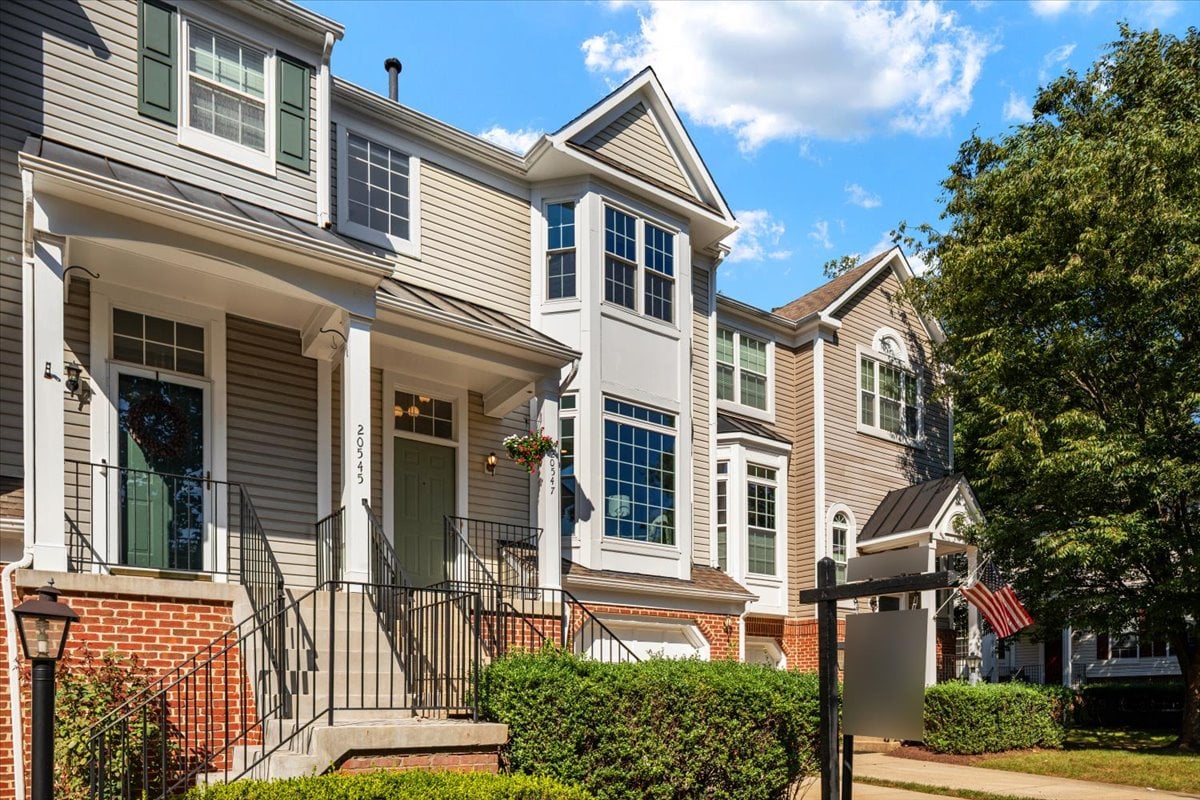 Attached townhomes with brick lower facades, front steps, and a property sign beside the sidewalk.