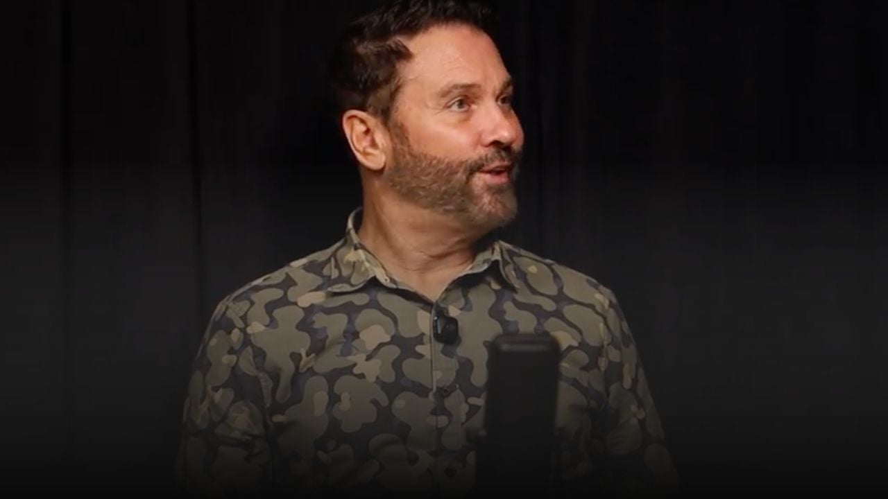 Man in a camouflage shirt speaking onstage with a microphone clipped to his shirt.