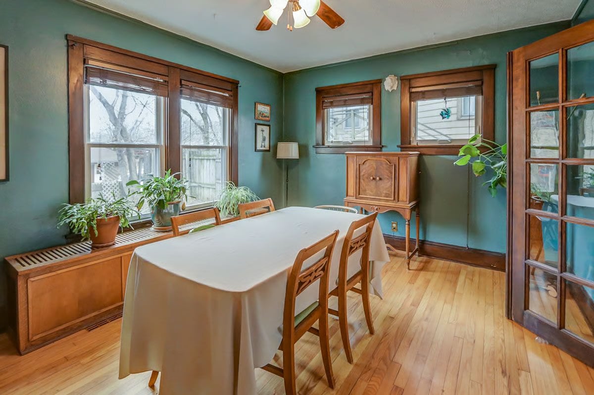 Dining room with a long table, teal walls, hardwood floors, and a window bench lined with plants.
