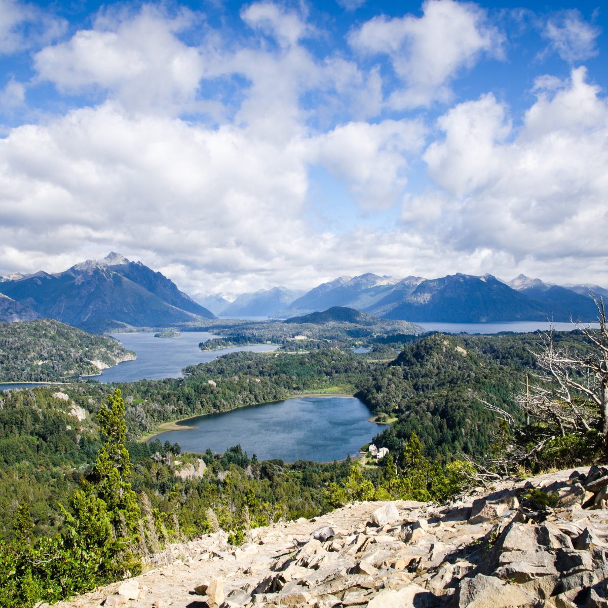 Mountain lakes wind through forested hills below a rocky overlook and cloudy blue sky.