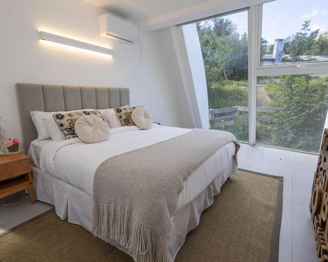 Staged bedroom with a white bed, gray upholstered headboard, and large windows overlooking greenery.