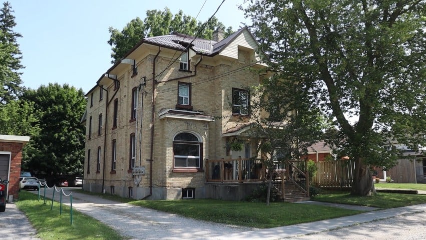 Large tan brick house with an arched window and raised porch beneath mature trees.