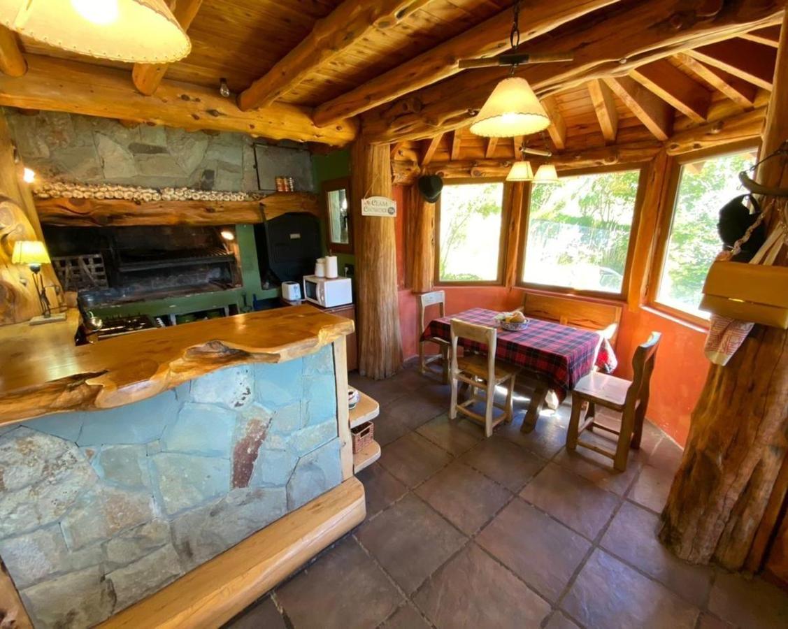 Rustic kitchen with exposed log beams, a stone fireplace and counter, and a plaid dining table by large windows.