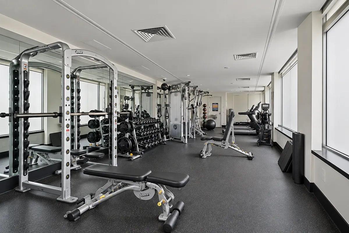 Building gym with mirrored walls, weight machines, dumbbell racks, and large windows.