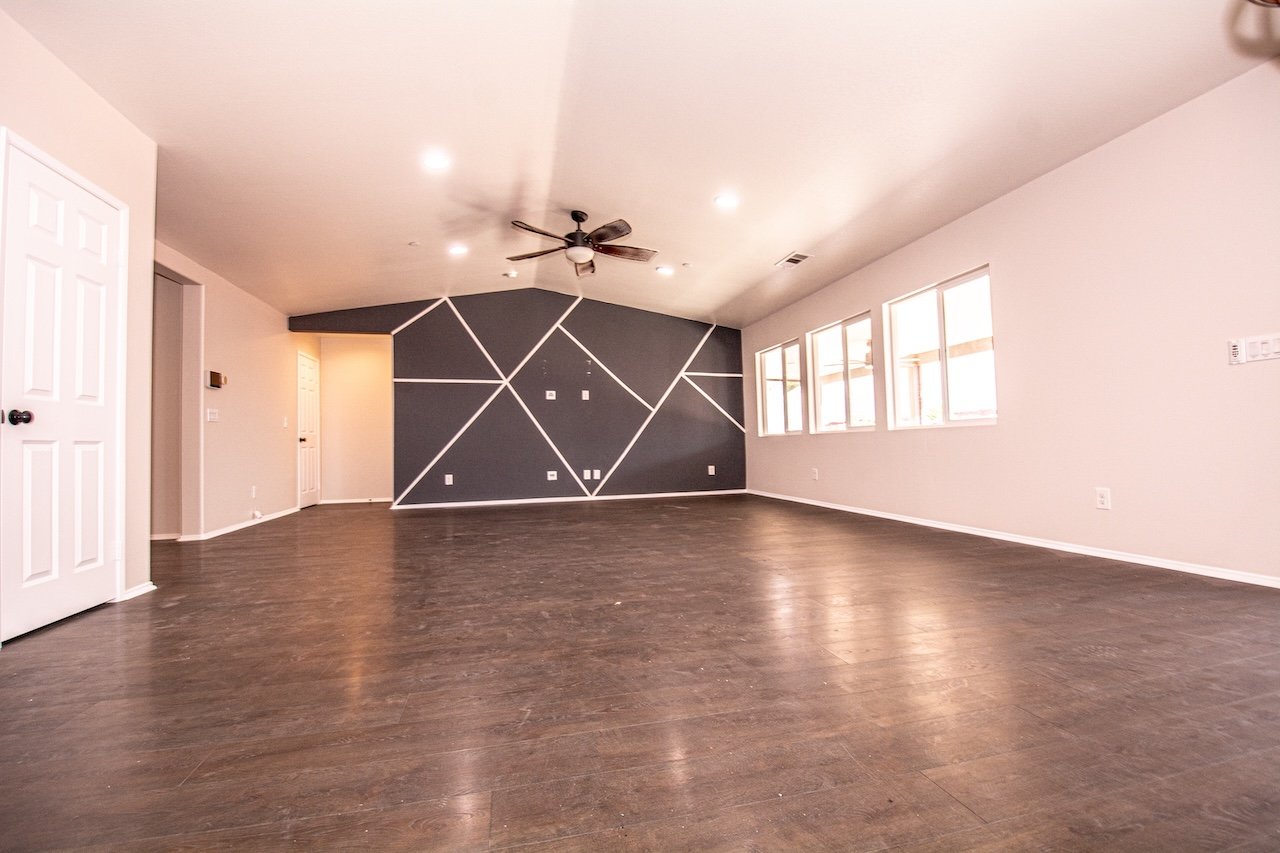 Vacant living room with dark wood floors, geometric accent wall, ceiling fan, and bright windows.