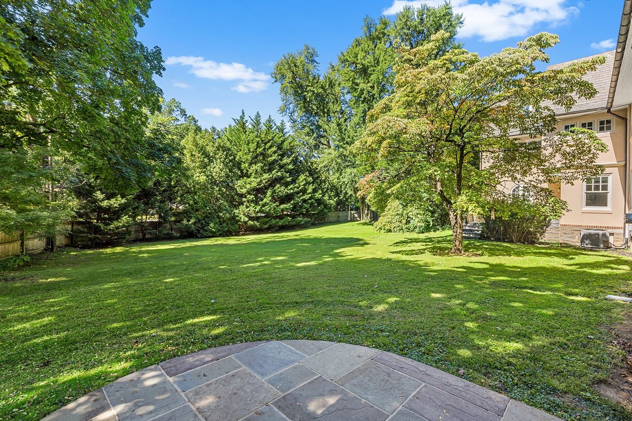 Spacious backyard with a broad lawn, mature trees, and a stone patio beside the house.