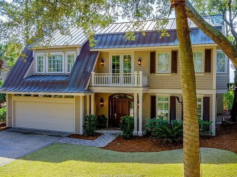 Two-story shingle home with a dark metal roof, attached garage, and upper balcony beneath mature trees.
