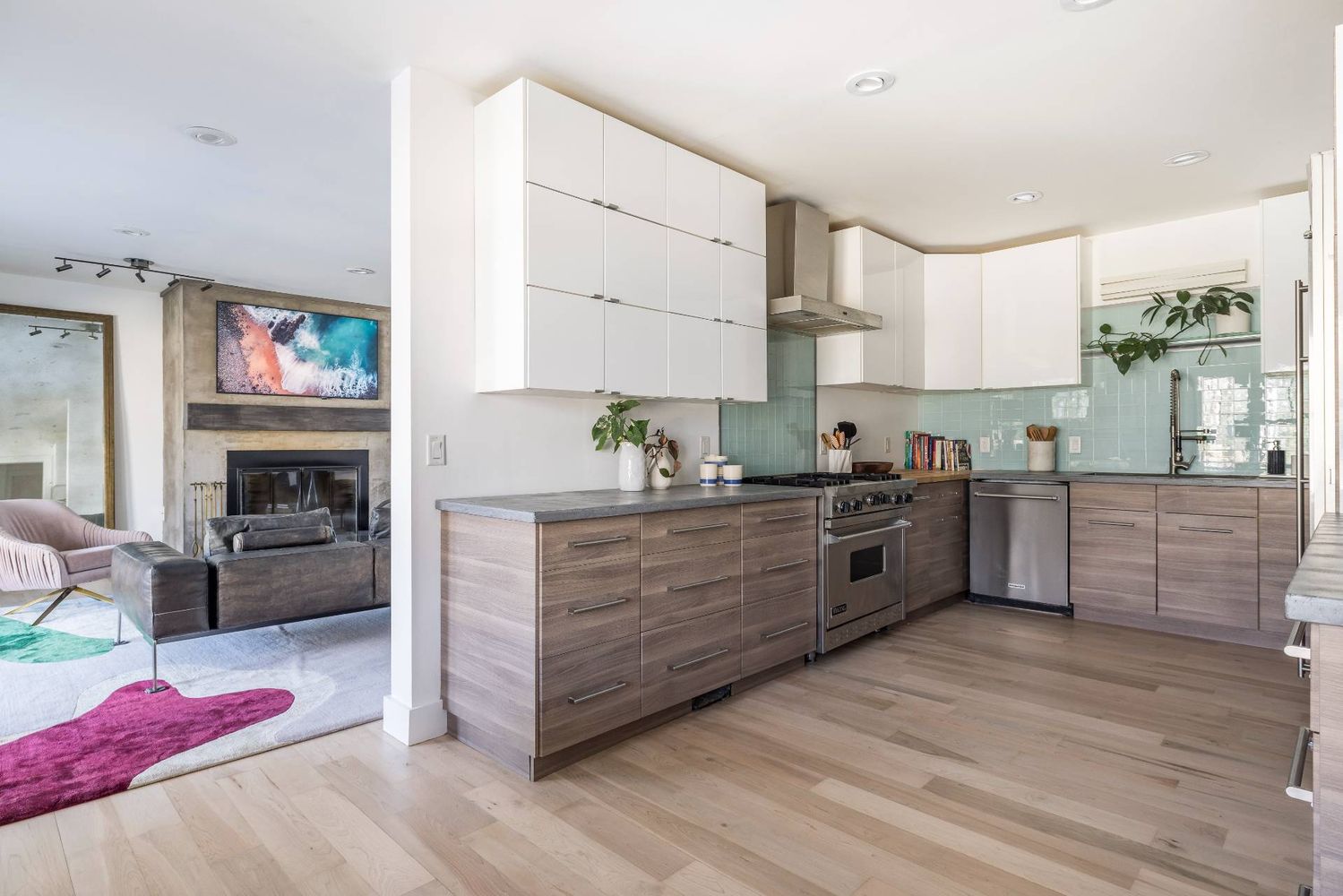 Open-plan kitchen with pale aqua tile backsplash and wood-tone cabinets beside a living room fireplace.