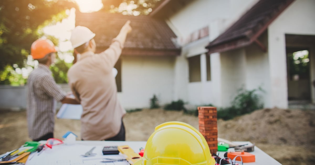 A table outside a newly-built home with architectural plans and supplies. In the background, two engineers discuss.