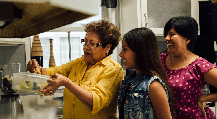 A woman in a yellow shirt cooks in a kitchen, watched by a smiling girl and another woman. The atmosphere is warm and cheerful, with a sense of family bonding.