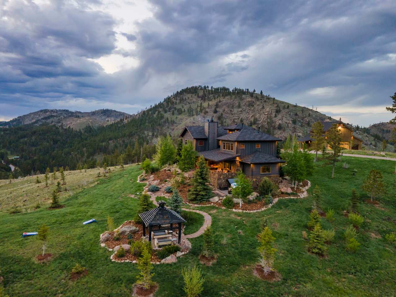 Mountain home on a landscaped hillside, with a gazebo and rocky ridges beneath dramatic clouds.