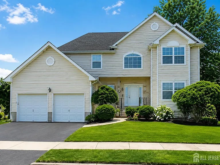 Two-story beige house with a two-car garage, landscaped shrubs, and a broad green lawn.