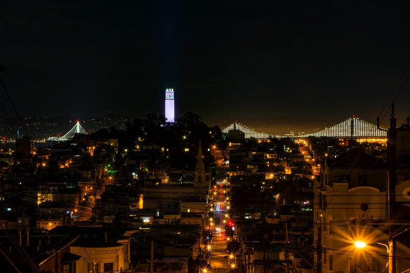 Nighttime view of San Francisco looking toward Telegraph Hill and Coit Tower, with the illuminated Bay Bridge stretching across the background and city streets glowing below.