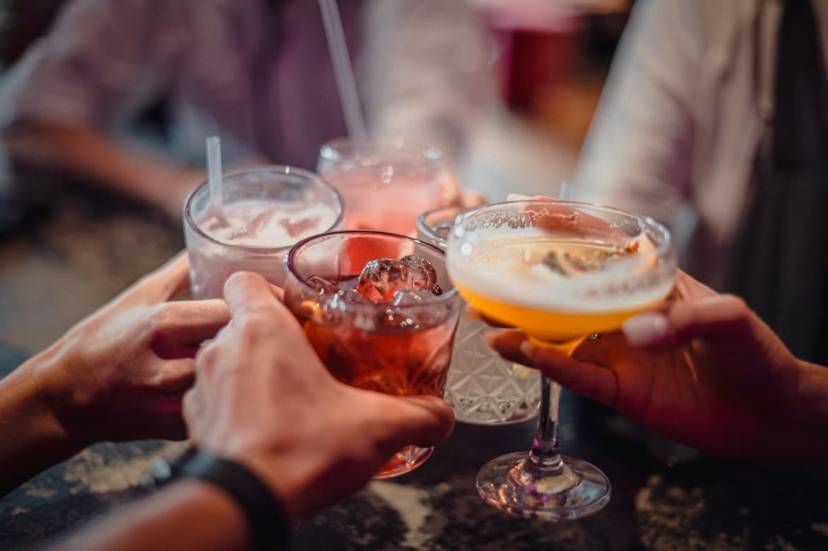 Friends raise cocktails together at a bar, including a beer and colorful mixed drinks.
