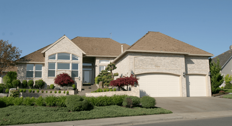 Single-story brick house with large arched windows, a two-car garage, and landscaped garden with trimmed bushes and red maple trees under a clear blue sky.