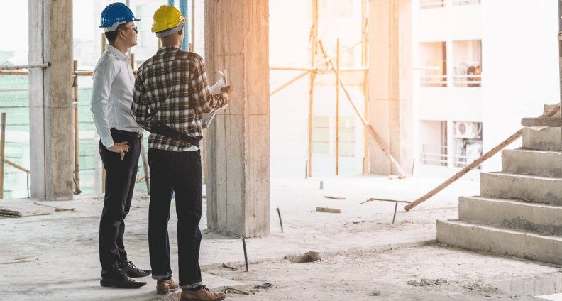 Unfinished building interior with two hard-hatted men inspecting concrete columns and stairs.