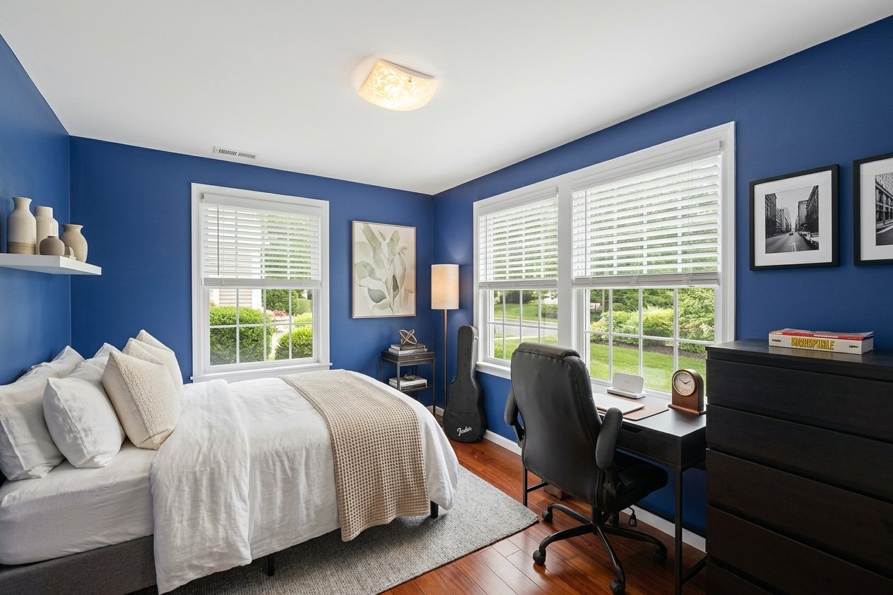 Bedroom with deep blue walls, a white bed, and a desk beside large windows.