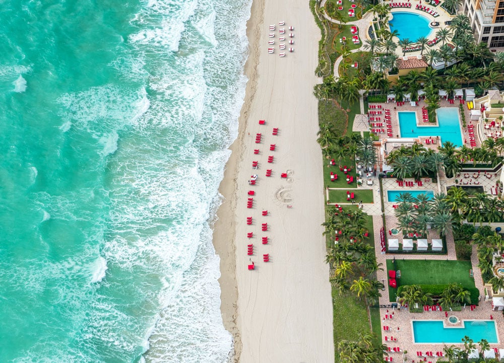 Aerial view of a beachfront resort with turquoise pools, palm trees, and red loungers along the shore.