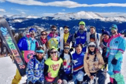Skiers in colorful winter gear pose with skis against a snowy mountain backdrop.